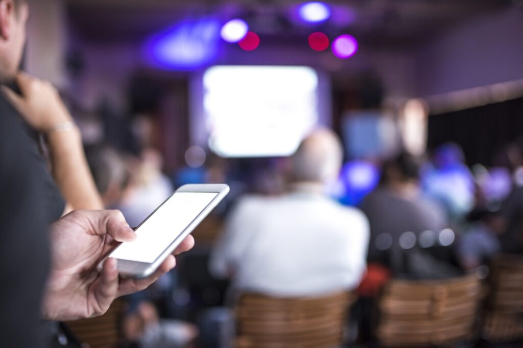 People sitting in a conference hall on a business seminar. Man's hand using mobile phone is in front, in focus, people defocused in back. There is white screen made by projector in front.