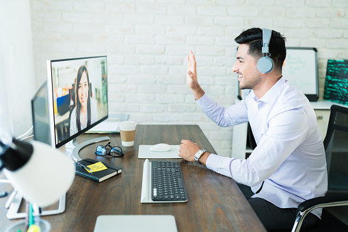 Confident young salesman waving at colleague during video call on computer at desk